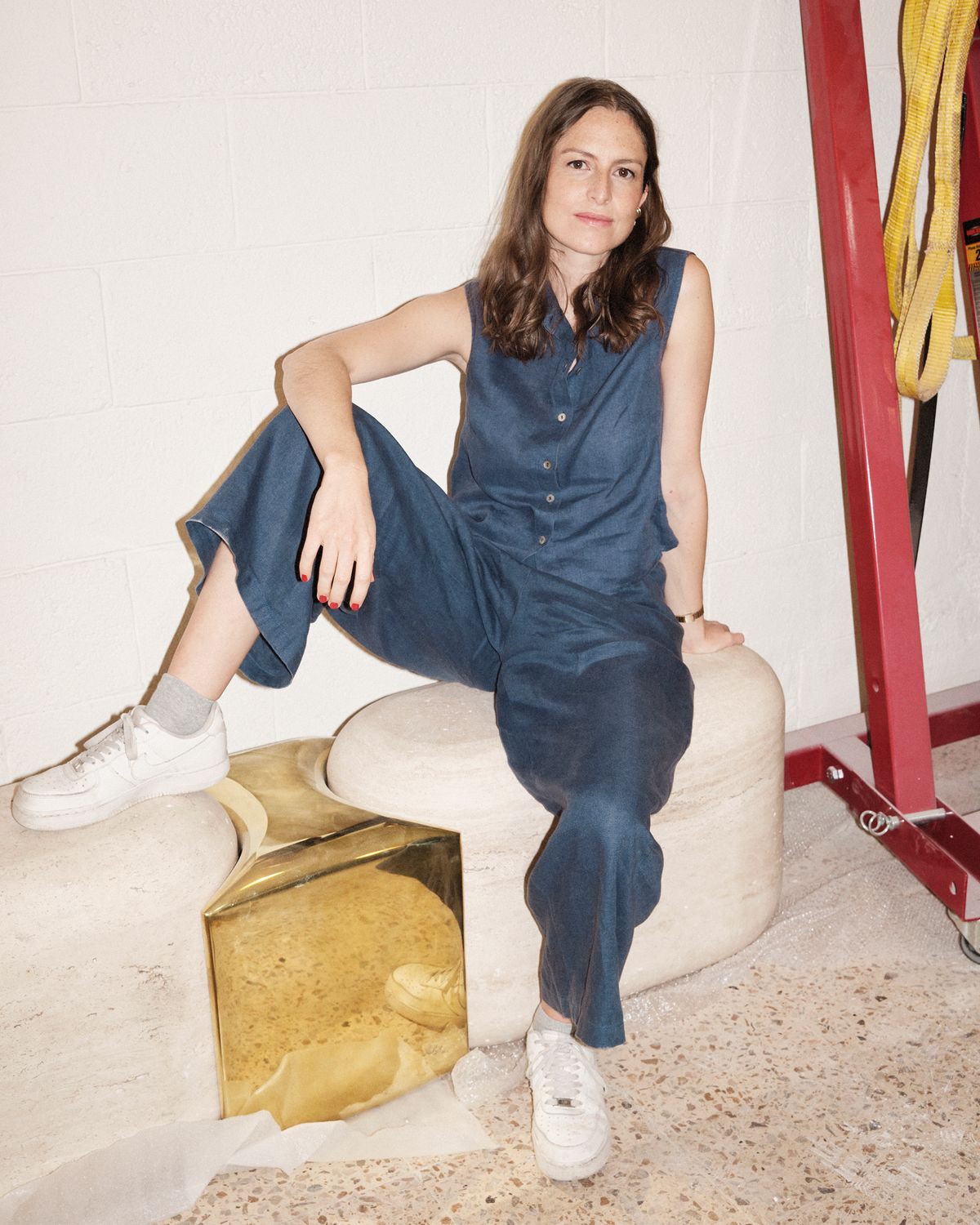 Artist Adeline de Monseignat in a navy linen jumpsuit and white sneakers, seated on a sculptural stone bench beside a gold metallic cube in a workshop