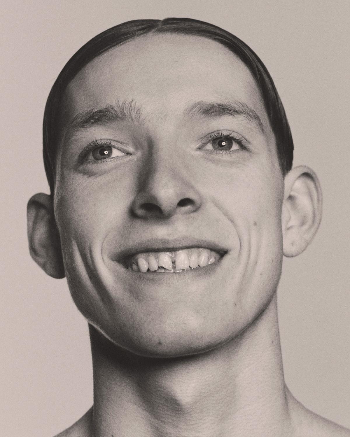 Sepia-toned close-up portrait of a smiling young man with a center-parted, slicked-back hairstyle, tilting his head and looking at the camera.