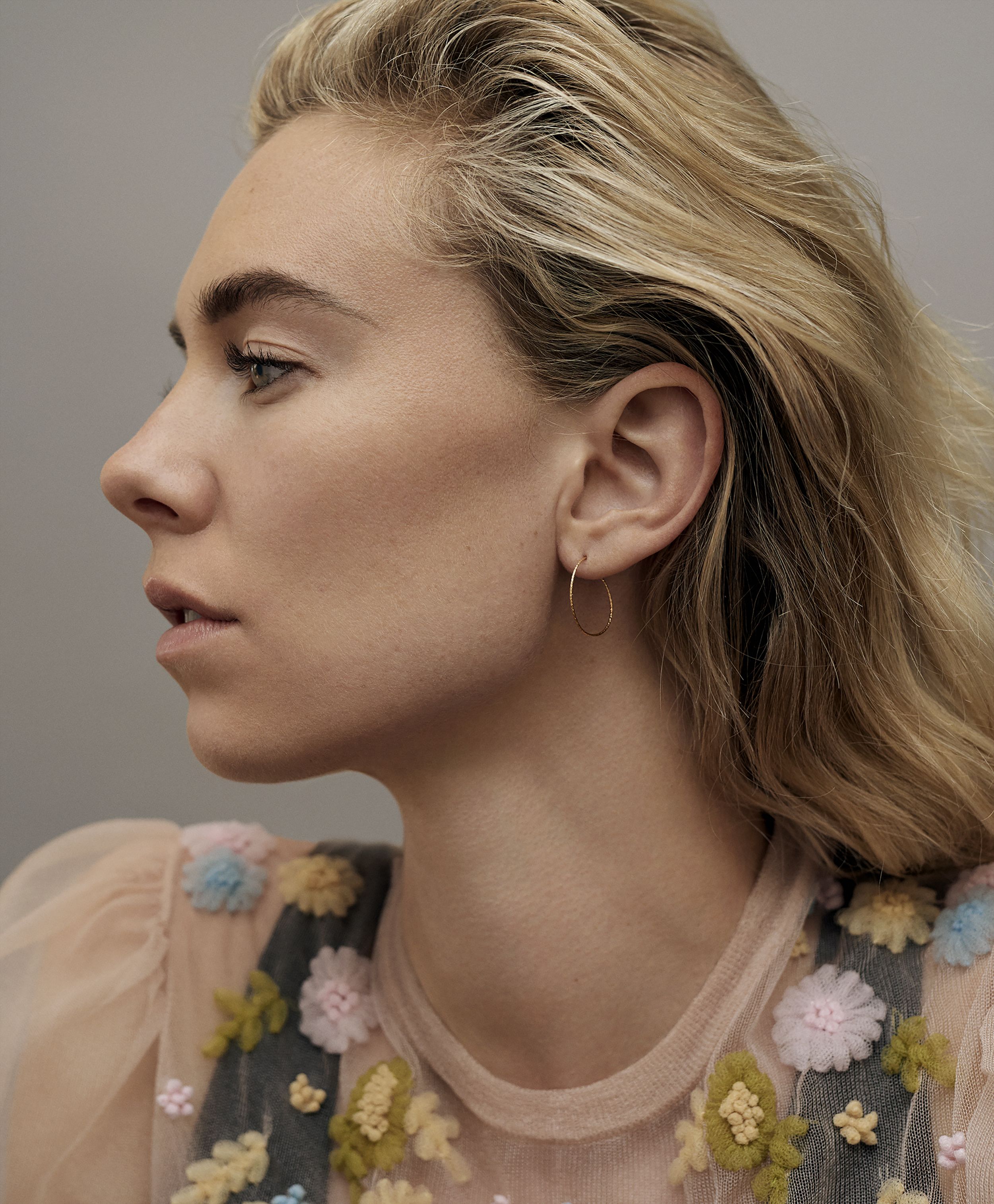 Side-profile portrait of actress Vanessa Kirby with swept-back blonde hair, a thin gold hoop earring, and a sheer peach top with pastel embroidered flowers.