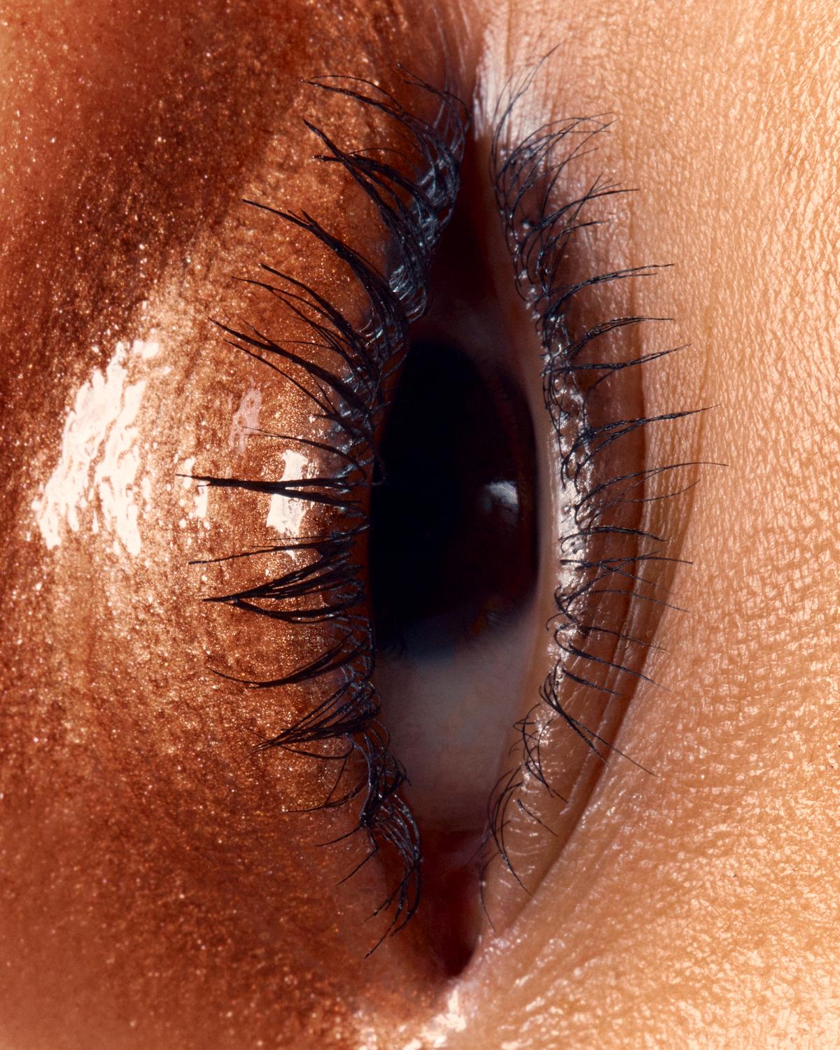Extreme close-up of an eye, shown sideways, with glossy bronze shimmer on the lid and defined black lashes.