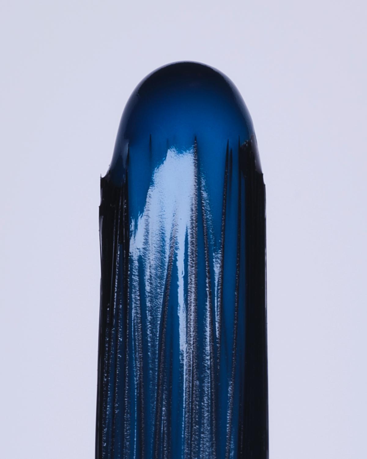 Macro still life of a nail varnish brush tip loaded with glossy, deep blue polish, with fine bristles and white highlights against a pale lavender background