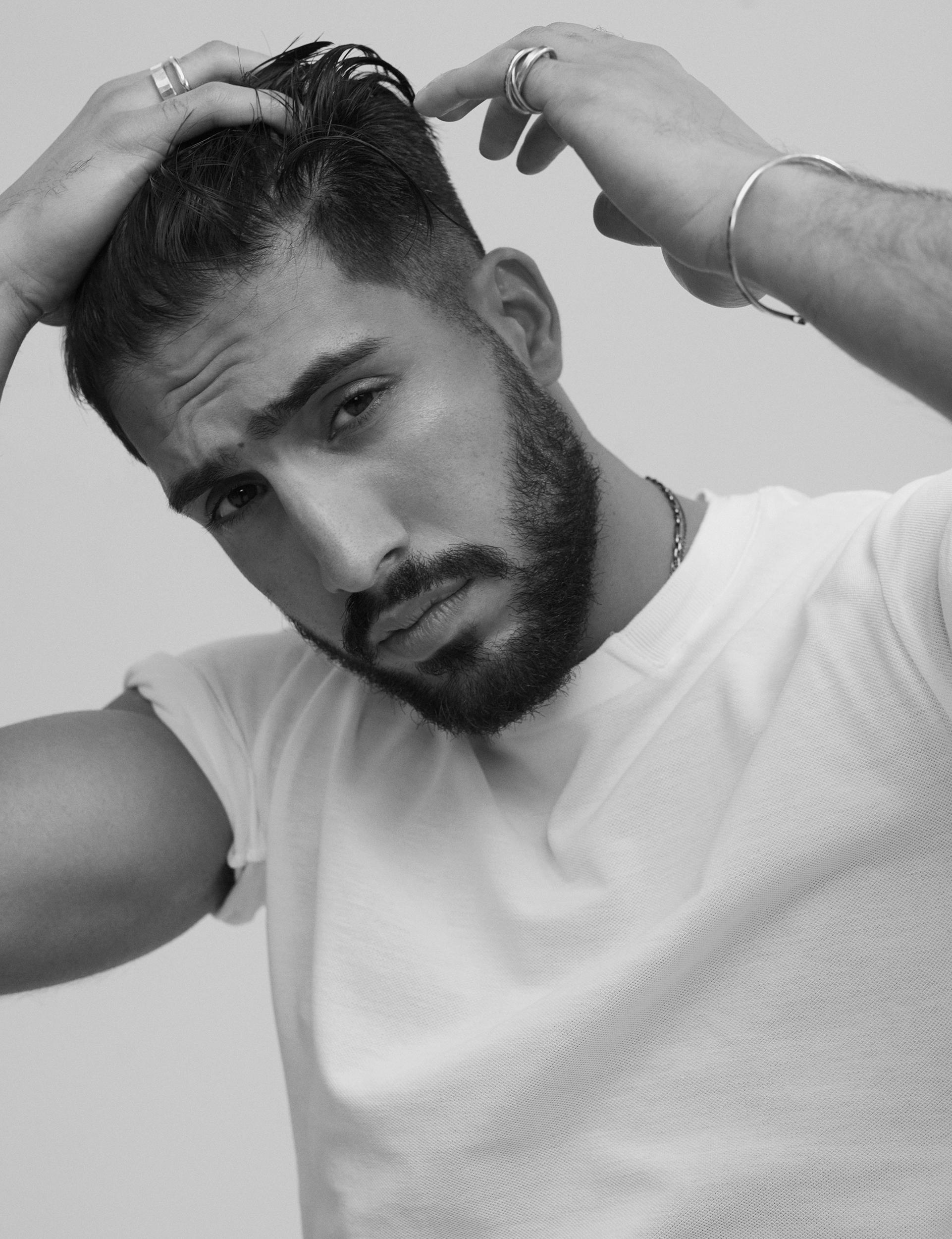 Black-and-white portrait of footballer Emre Can in a white tee and silver rings, with both hands in his swept-back hair, looking at the camera.