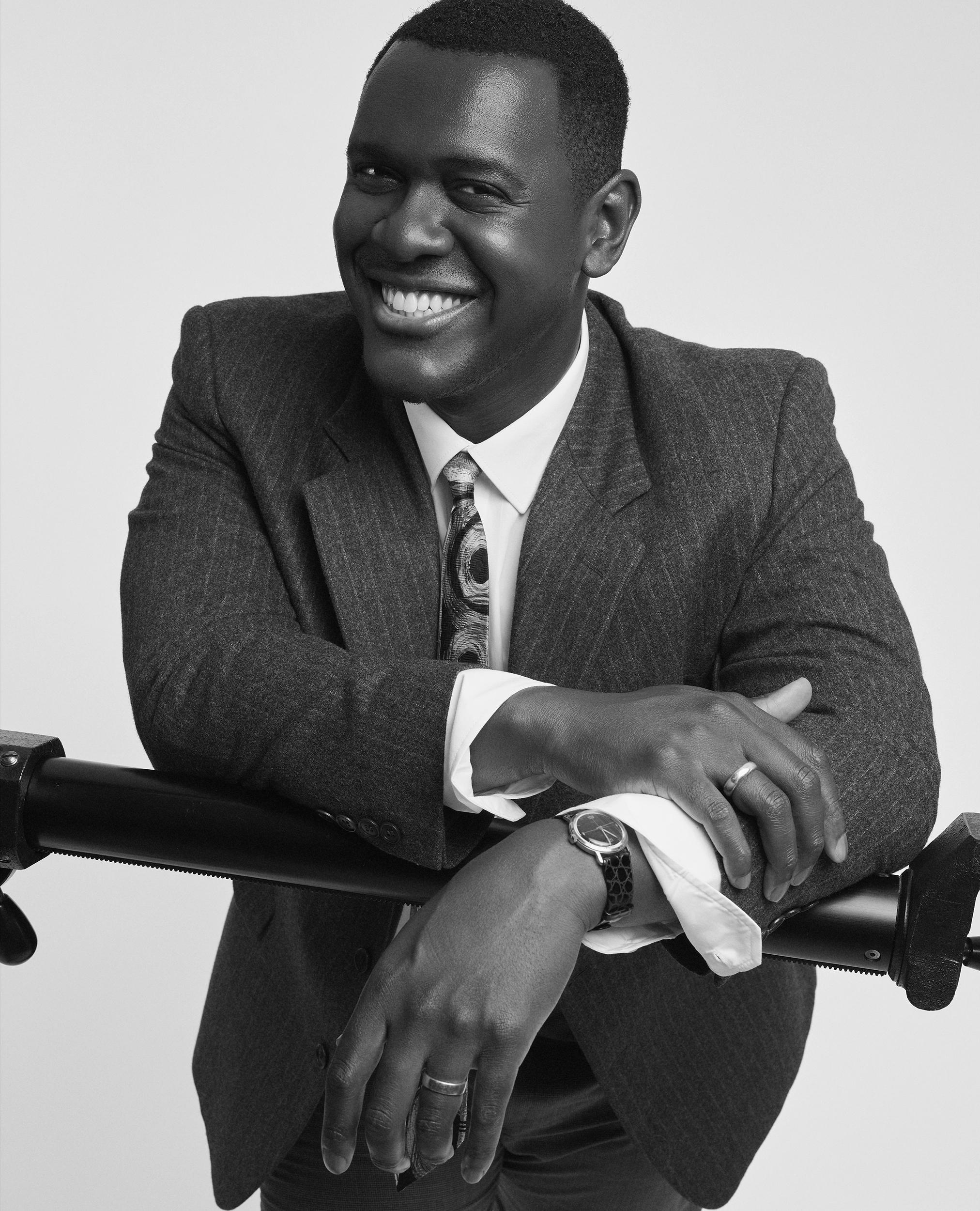 Black-and-white portrait of Abubakar Salim smiling in a pinstripe jacket and patterned tie, leaning his folded arms on a black bar.