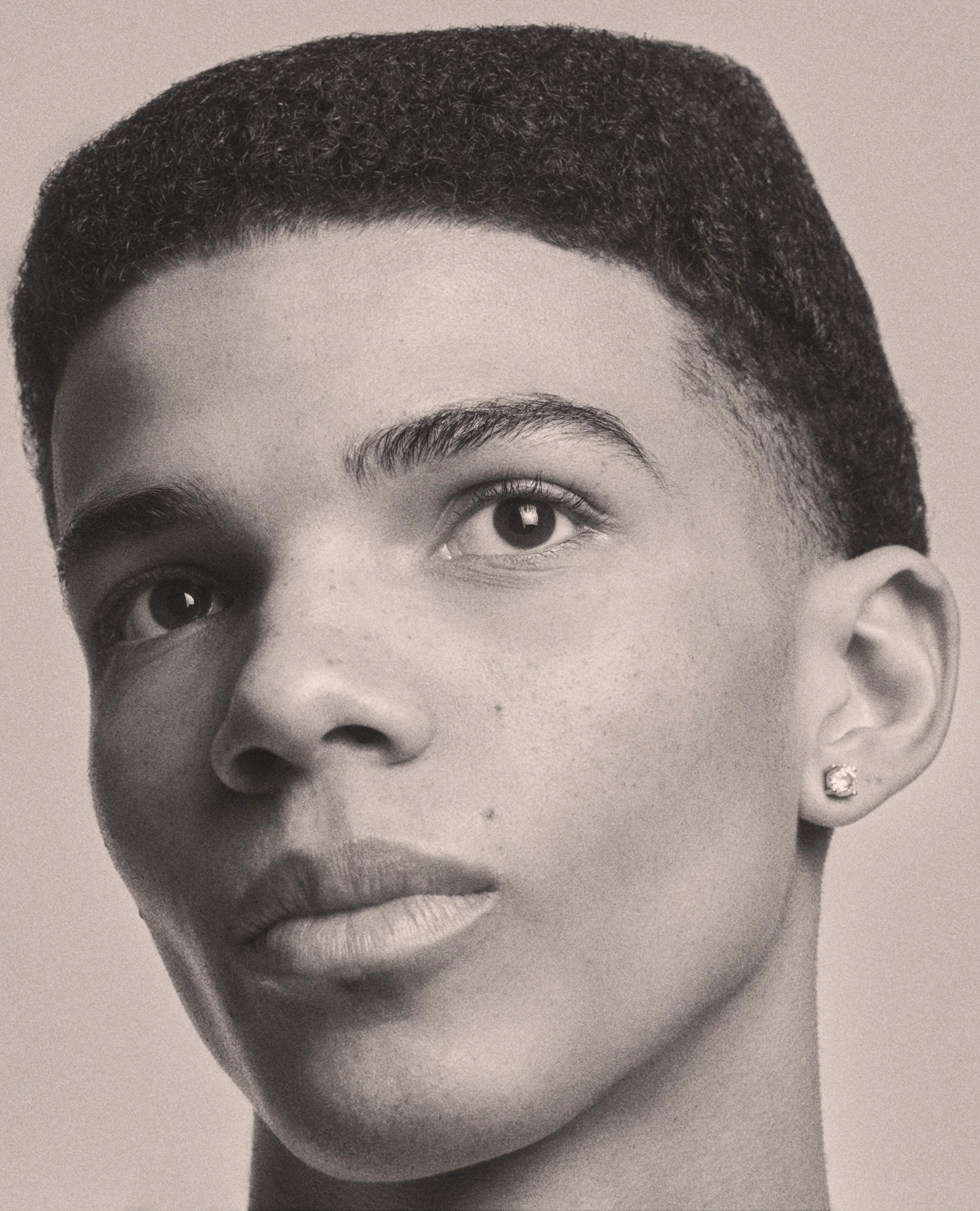 Sepia-toned close-up portrait of a young man with a short, flat-top fade and a diamond stud earring, his head tilted as he looks toward the camera.