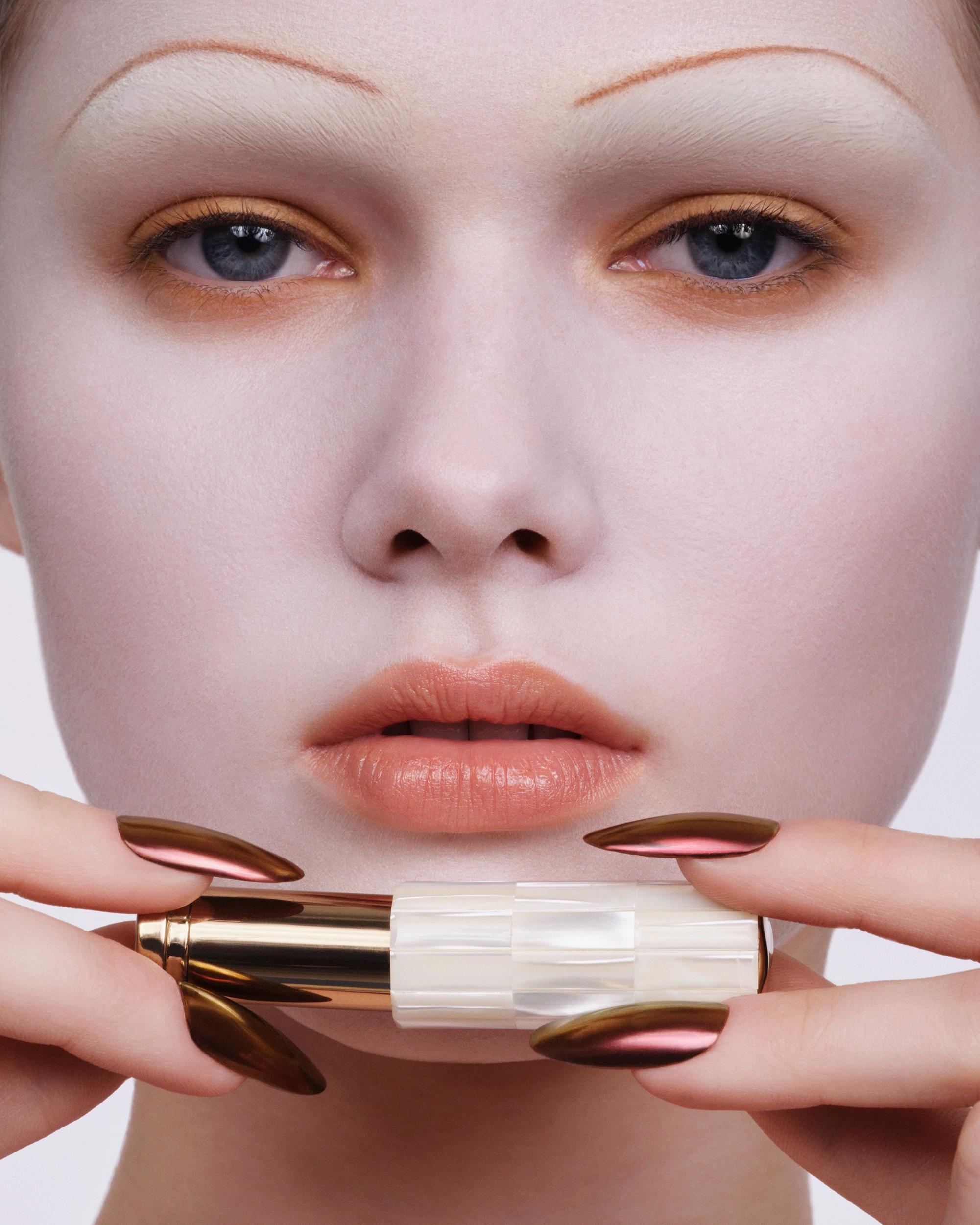 Close-up of a woman with a matte porcelain-white complexion, amber eyeshadow, and peach lips, holding a gold and pearl lipstick with chrome bronze nails