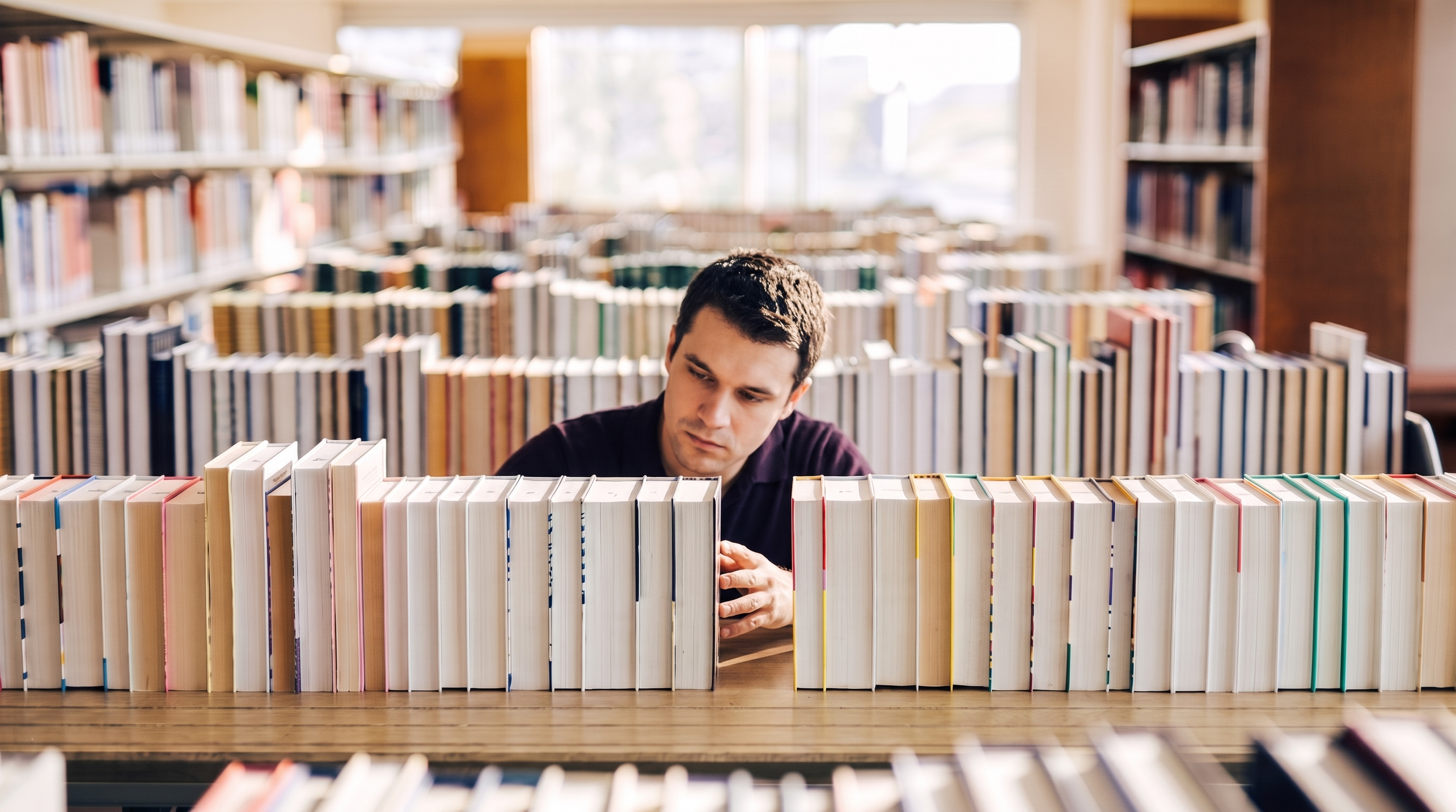 A man faces the camera while reaching for a book, eyes down on the spines beside him as he weighs the choice.