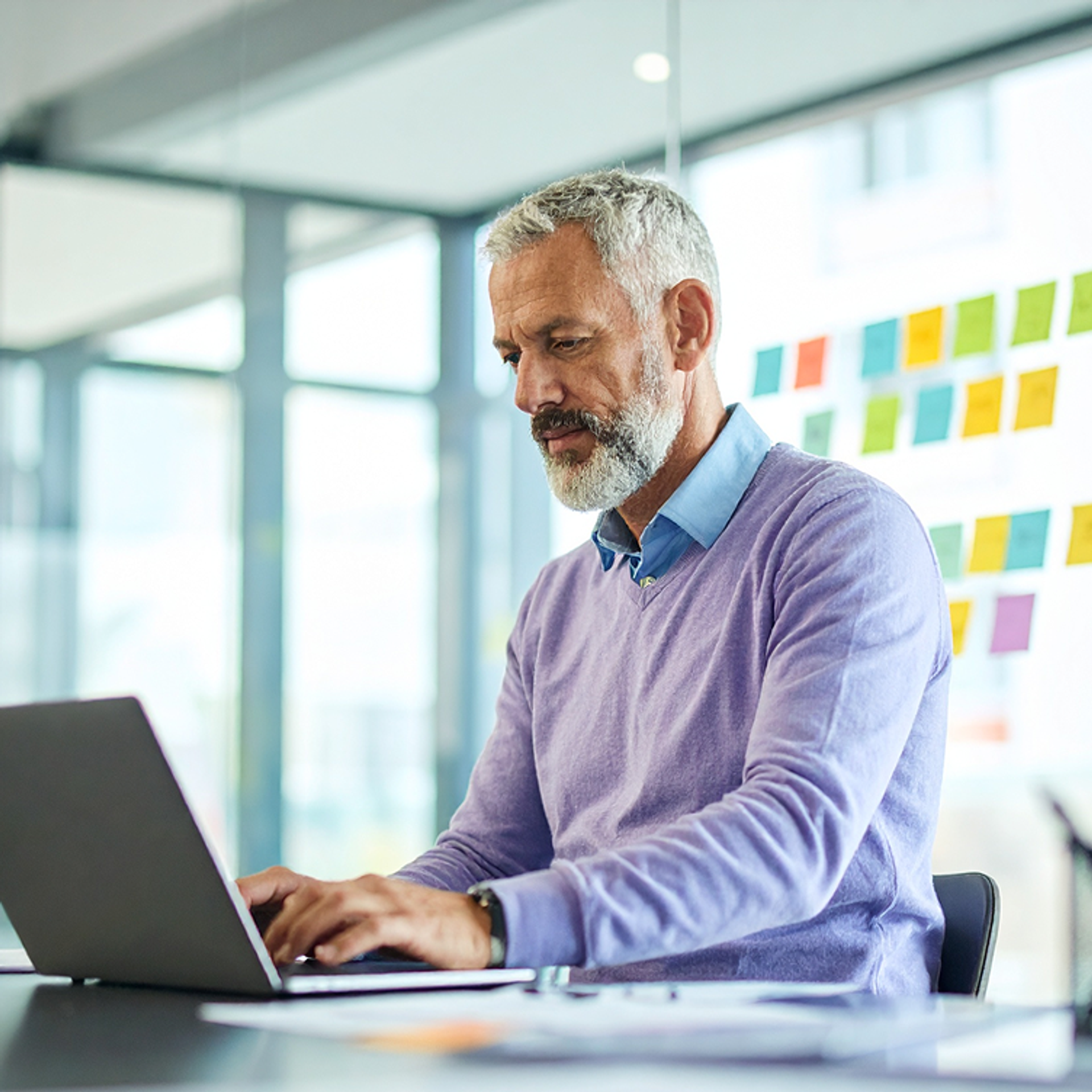 Man interacting with an AI chatbot on a laptop in a modern office, with colorful sticky notes on a glass wall and papers on the desk.