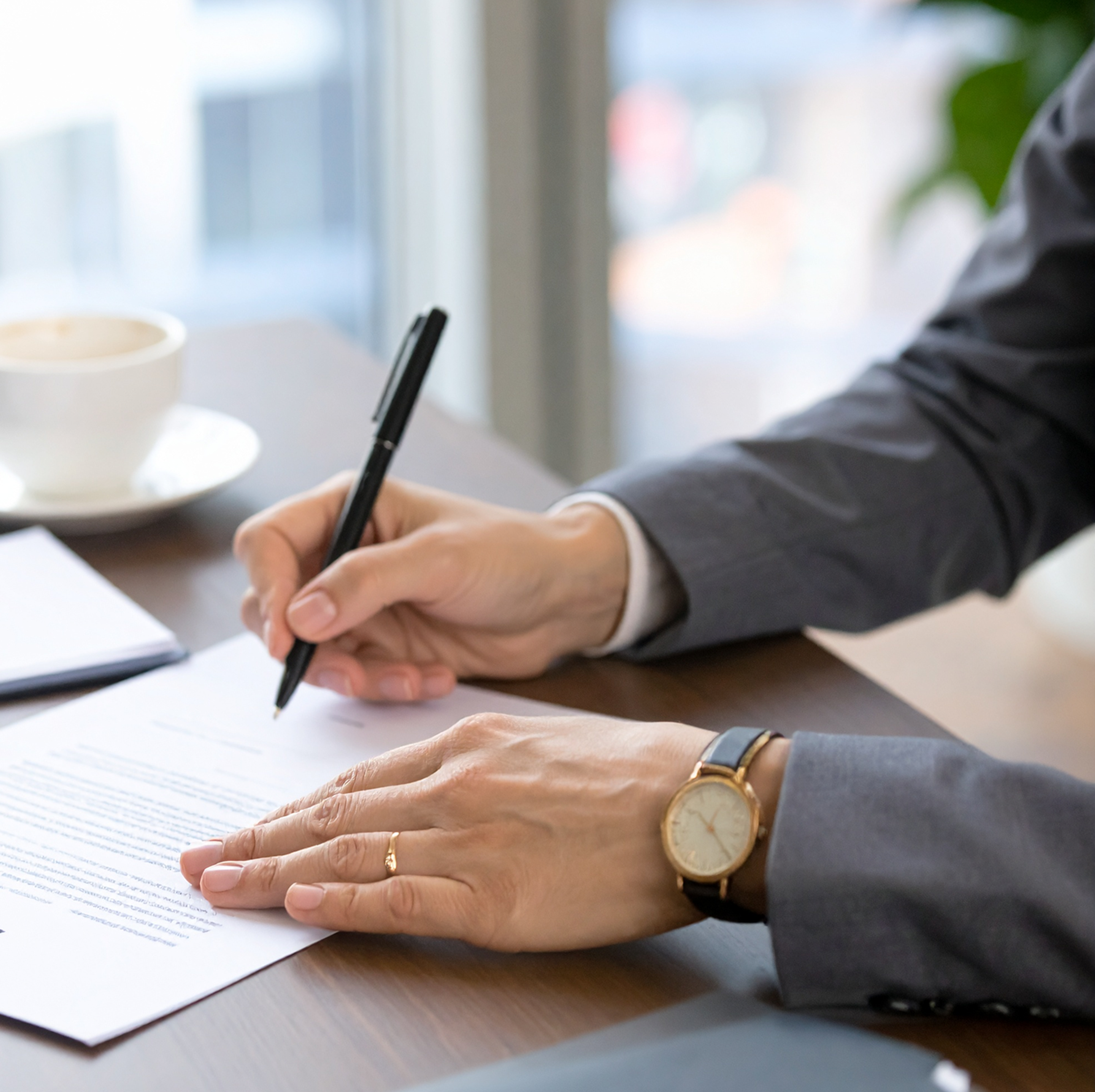 A person in a grey business suit signing a legal contract at an office desk.