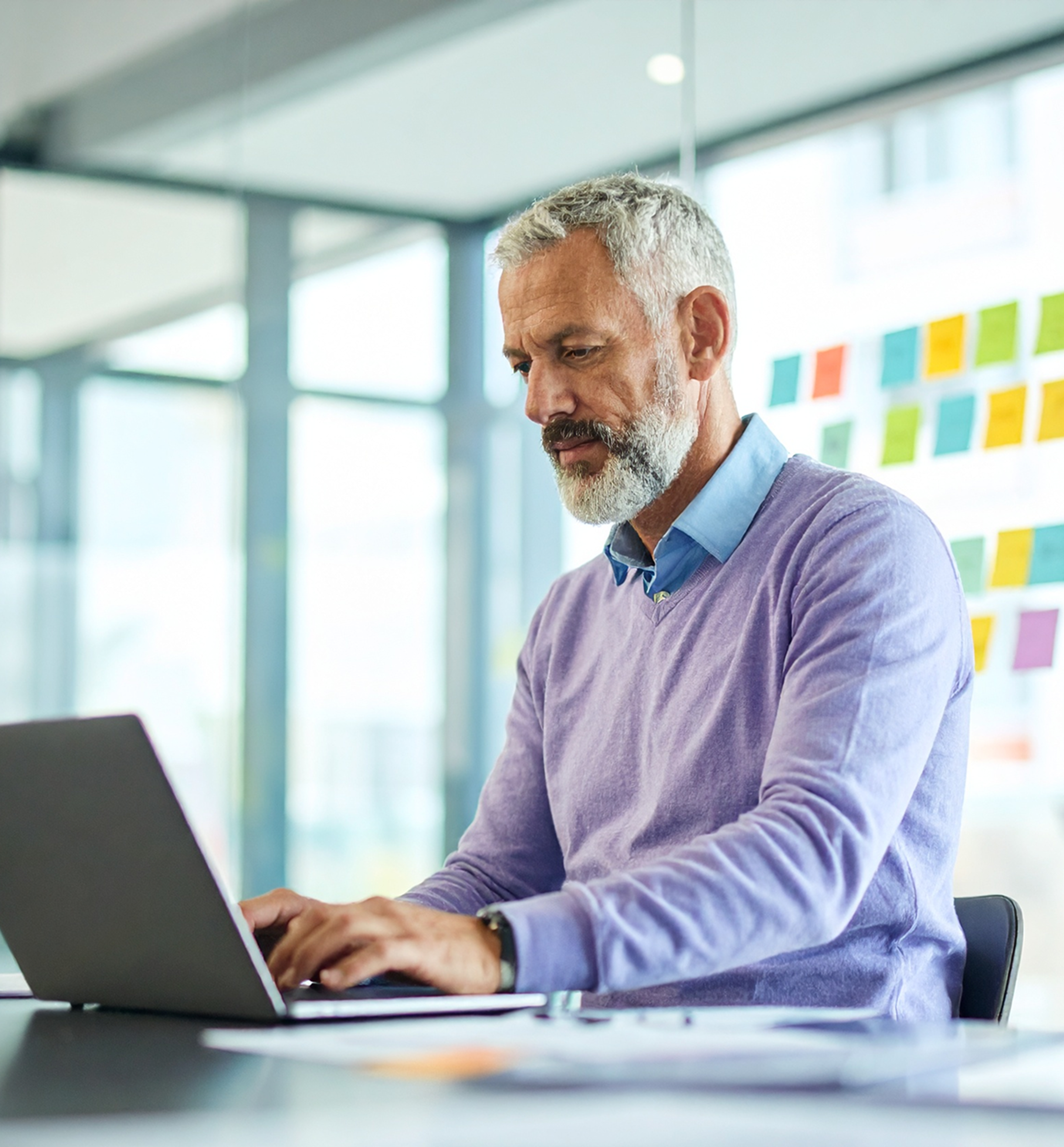 Man interacting with an AI chatbot on a laptop in a modern office, with colorful sticky notes on a glass wall and papers on the desk.