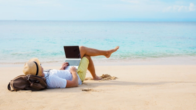 A men laying on the beach, with his head on his backpack and his laptop on his legs.