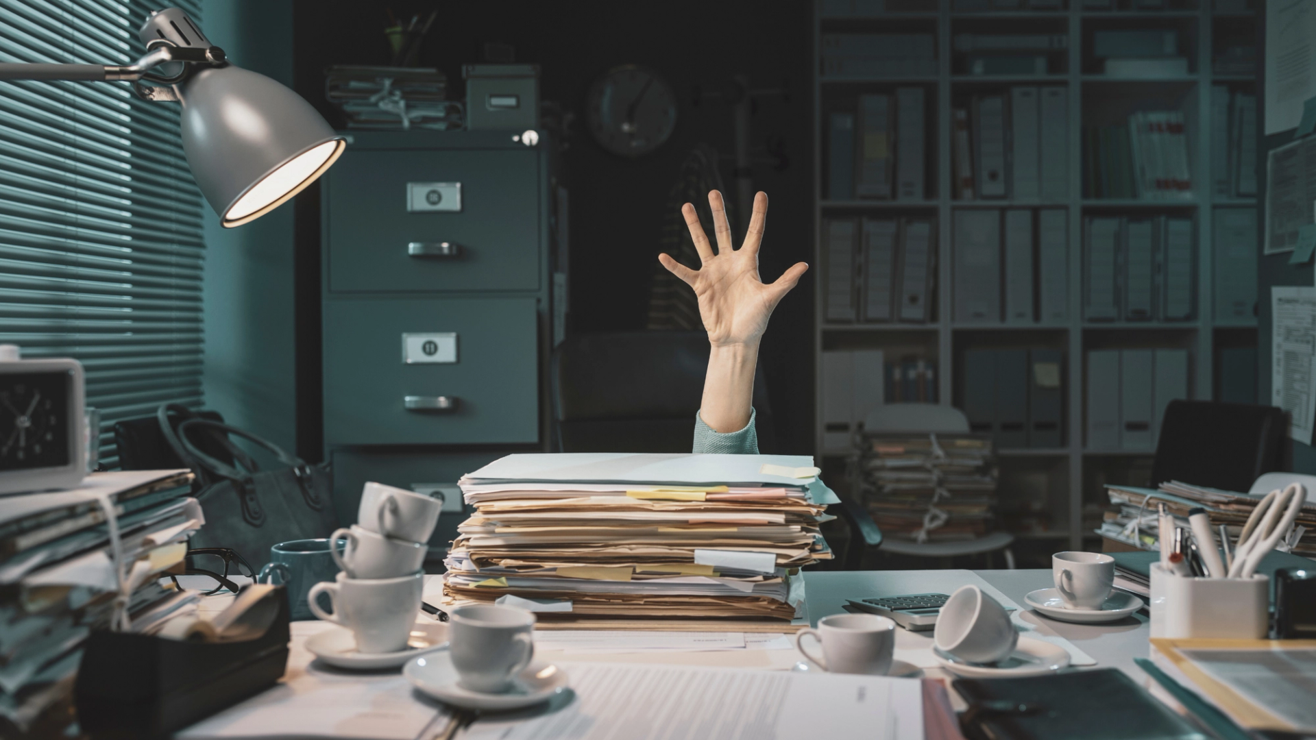 Hand asking for help on a desk full of papers and coffee cups