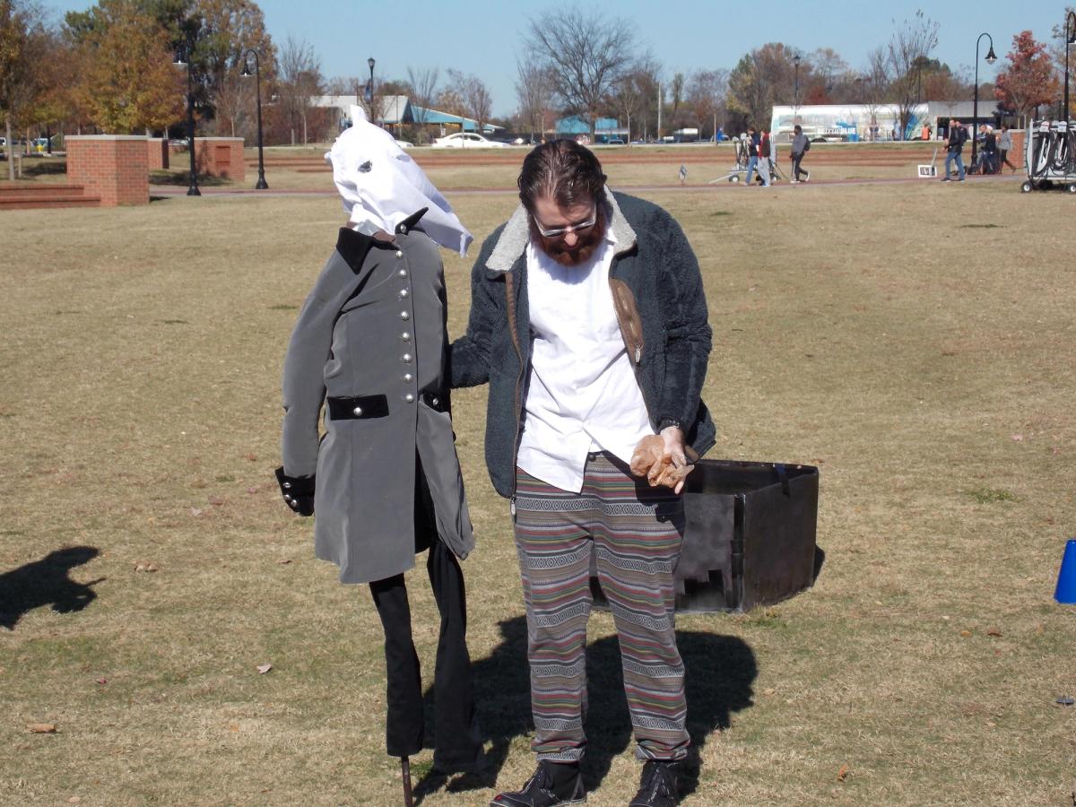 A student stands on a lawn beside an effigy of Forrest wearing a gray Confederate coat and a white hood, with a black coffin behind them.