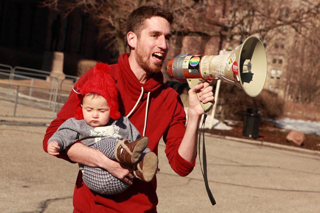 A striker in a red hoodie carries a baby in a sling in one arm and shouts into a sticker-covered megaphone held in the other.