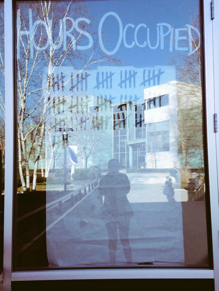A window of the occupied building with "Hours occupied" written across the top and rows of tally marks below, reflecting the campus outside and the silhouette of the photographer.