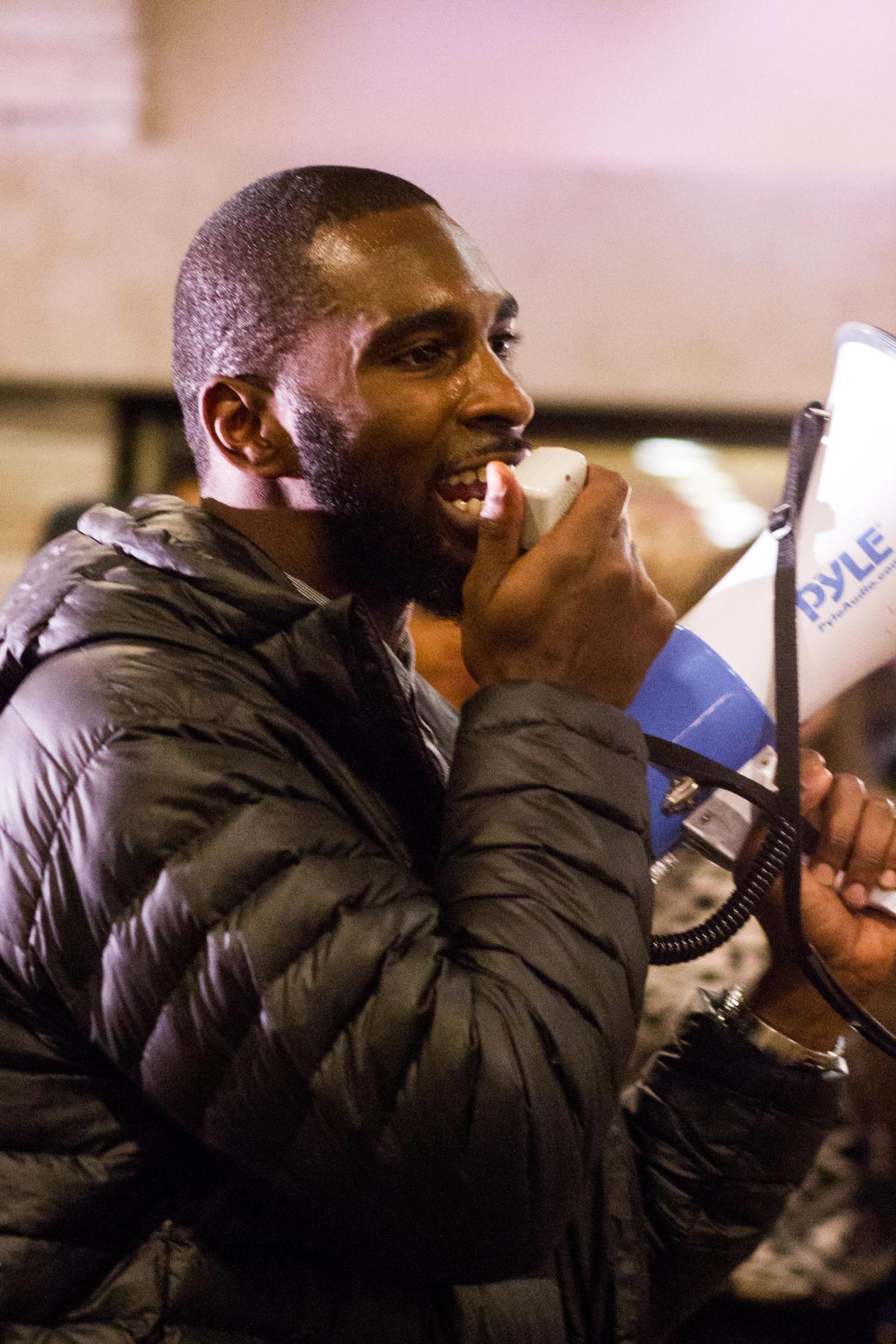 A Black man in a black puffer jacket shouts into a white and blue megaphone.