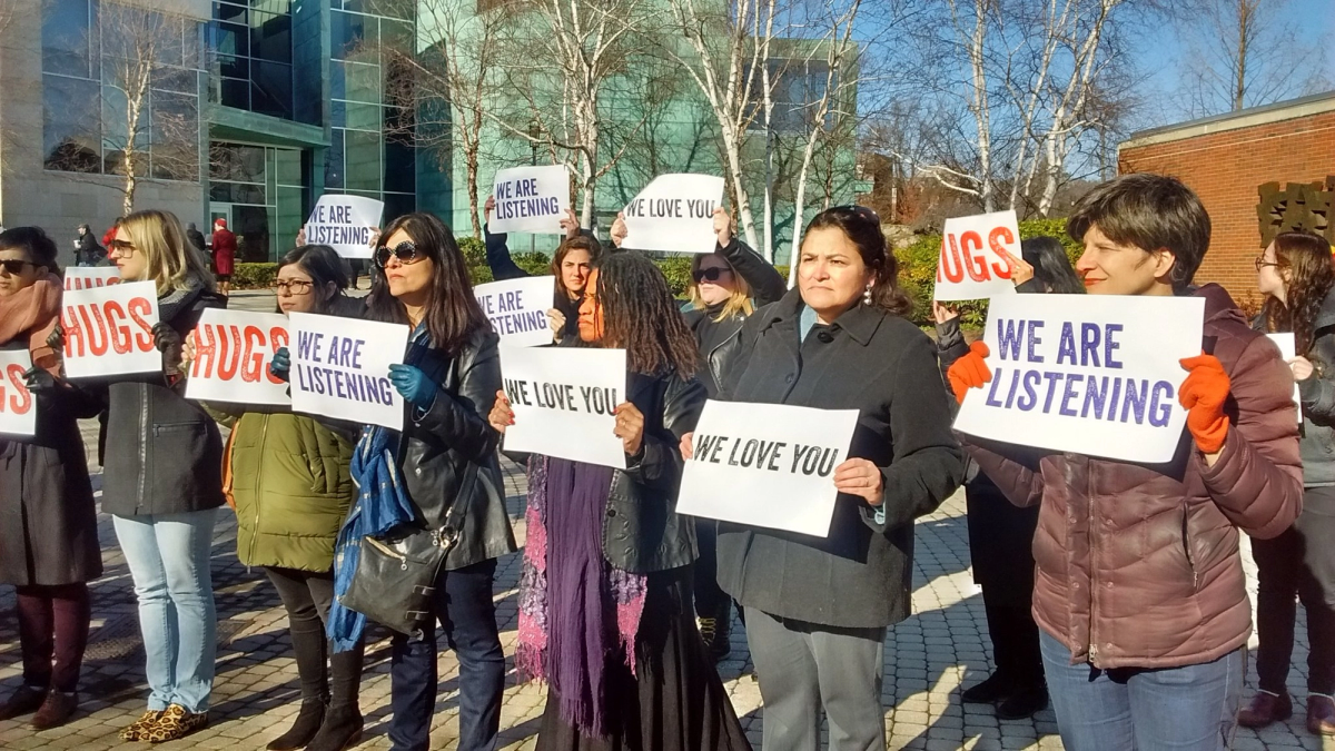 A line of faculty members outdoors on a sunny day hold printed signs reading "We are listening," "We love you," and "Hugs."
