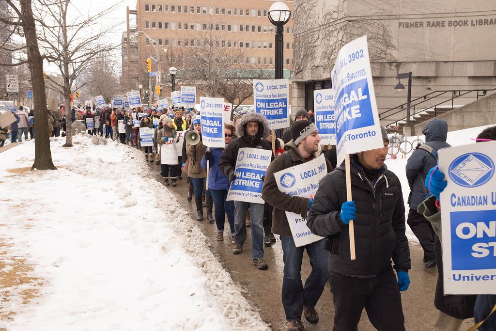 A long line of picketers in winter coats carry blue “Local 3902 — On Legal Strike” placards along a snowy sidewalk beside the Fisher Rare Book Library.