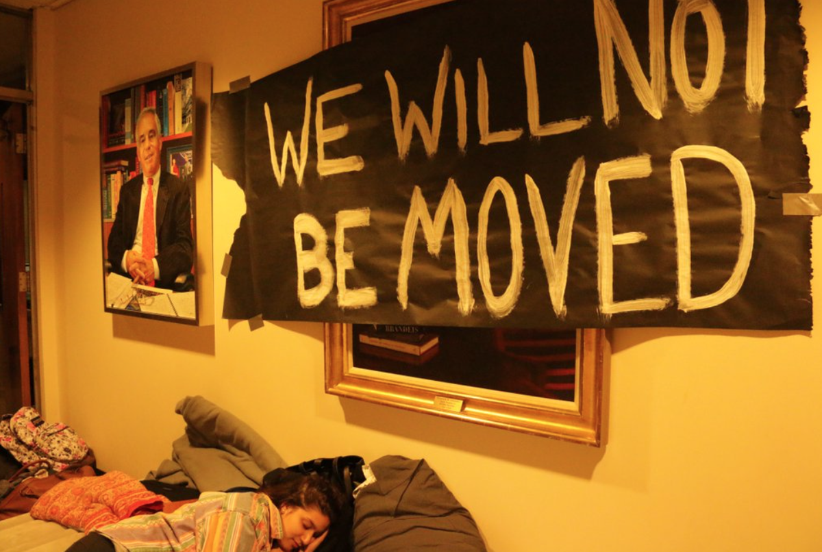 A student sleeps on blankets on the floor of an administrative office beneath a hand-painted black banner reading "We will not be moved," taped over framed portraits.