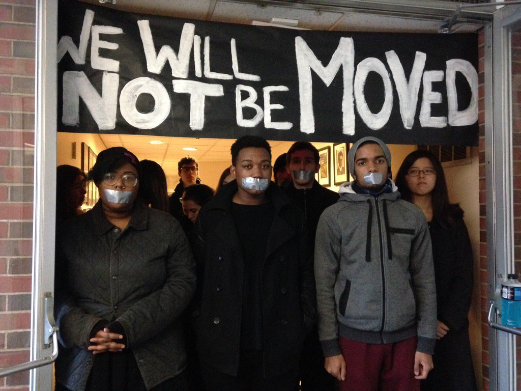 Students with tape over their mouths stand silently in a doorway beneath a hand-painted black banner reading "We will not be moved."