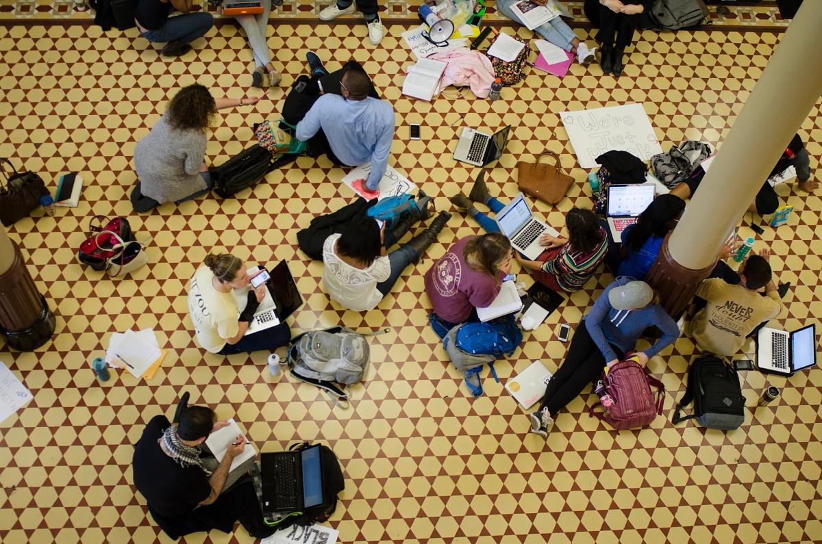 Seen from above, students sit on the tiled floor of Jesse Hall with laptops, books, and backpacks, alongside hand-lettered signs and a megaphone.