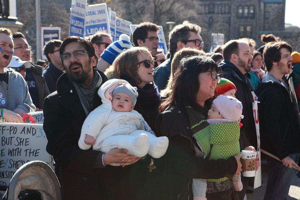 Parents on a picket line chant while holding infants in their arms and in a carrier, with CUPE 3902 “On Legal Strike” placards behind them.