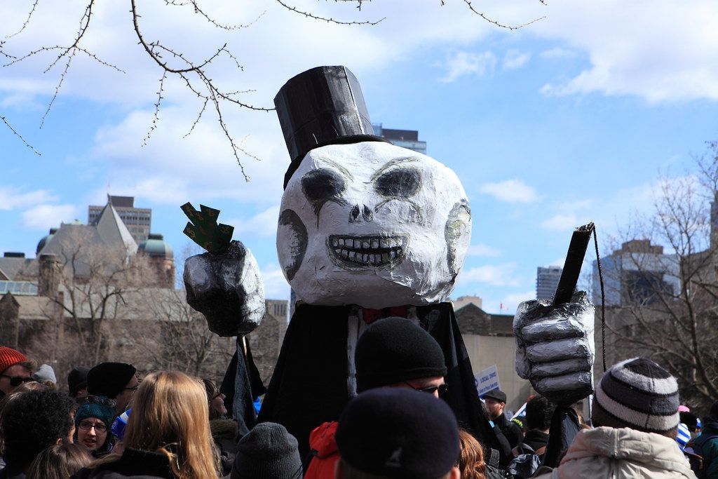 A giant papier-mâché puppet of a top-hatted skeletal figure towers over a crowd of picketers under a blue sky.