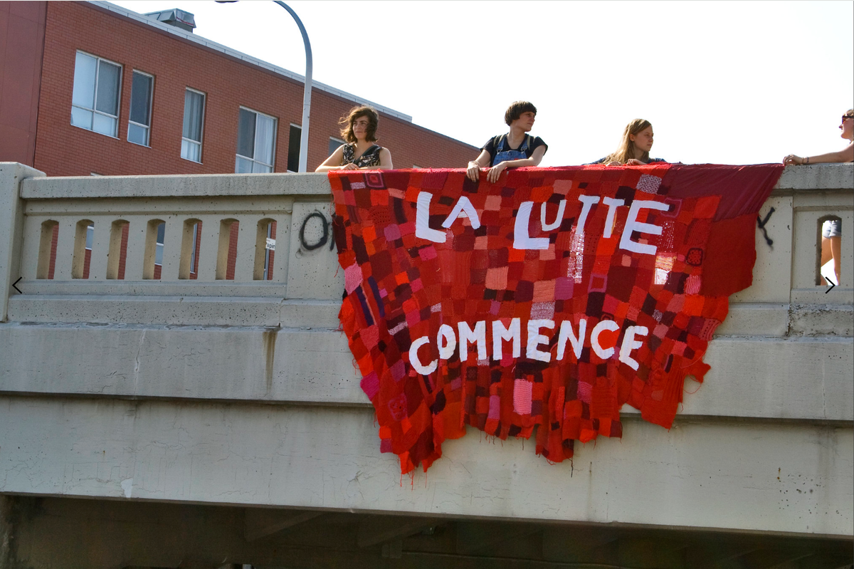 Four young people lean over a concrete overpass railing beside a huge patchwork banner of knitted red squares reading "La lutte commence."