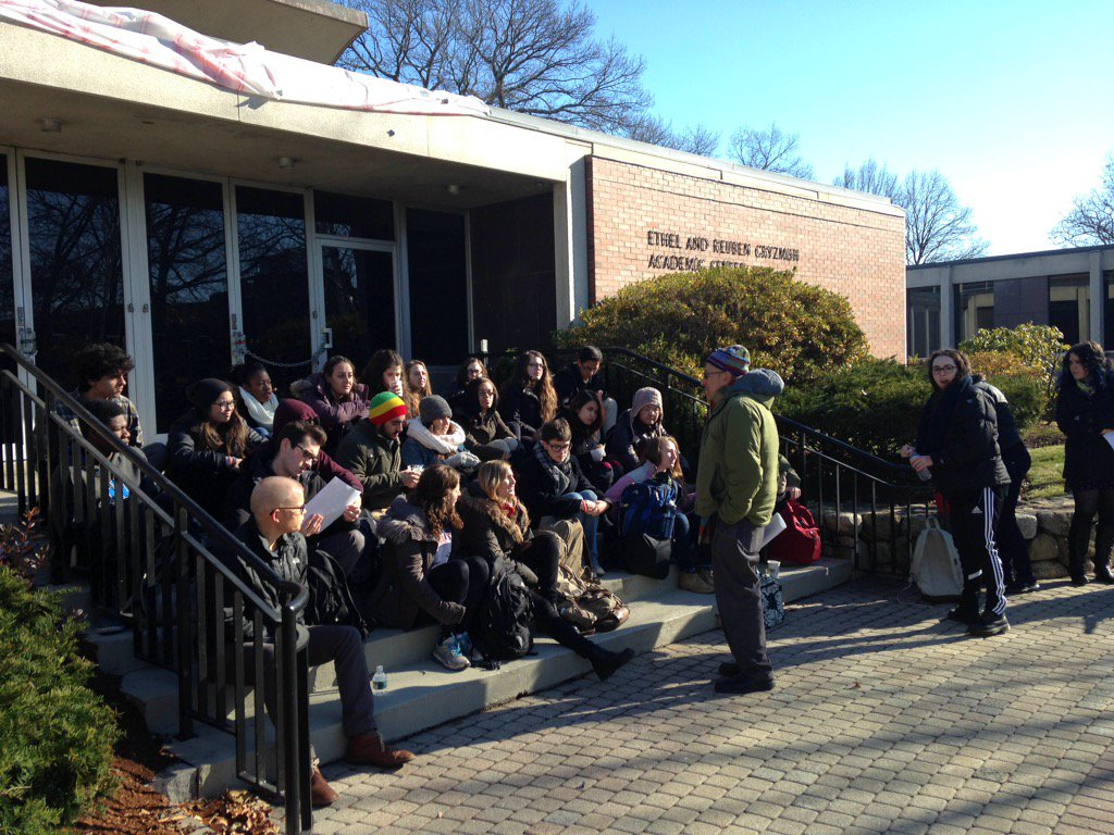 Students in winter coats sit on the steps of a brick building as an instructor in a green jacket and knit hat addresses them.