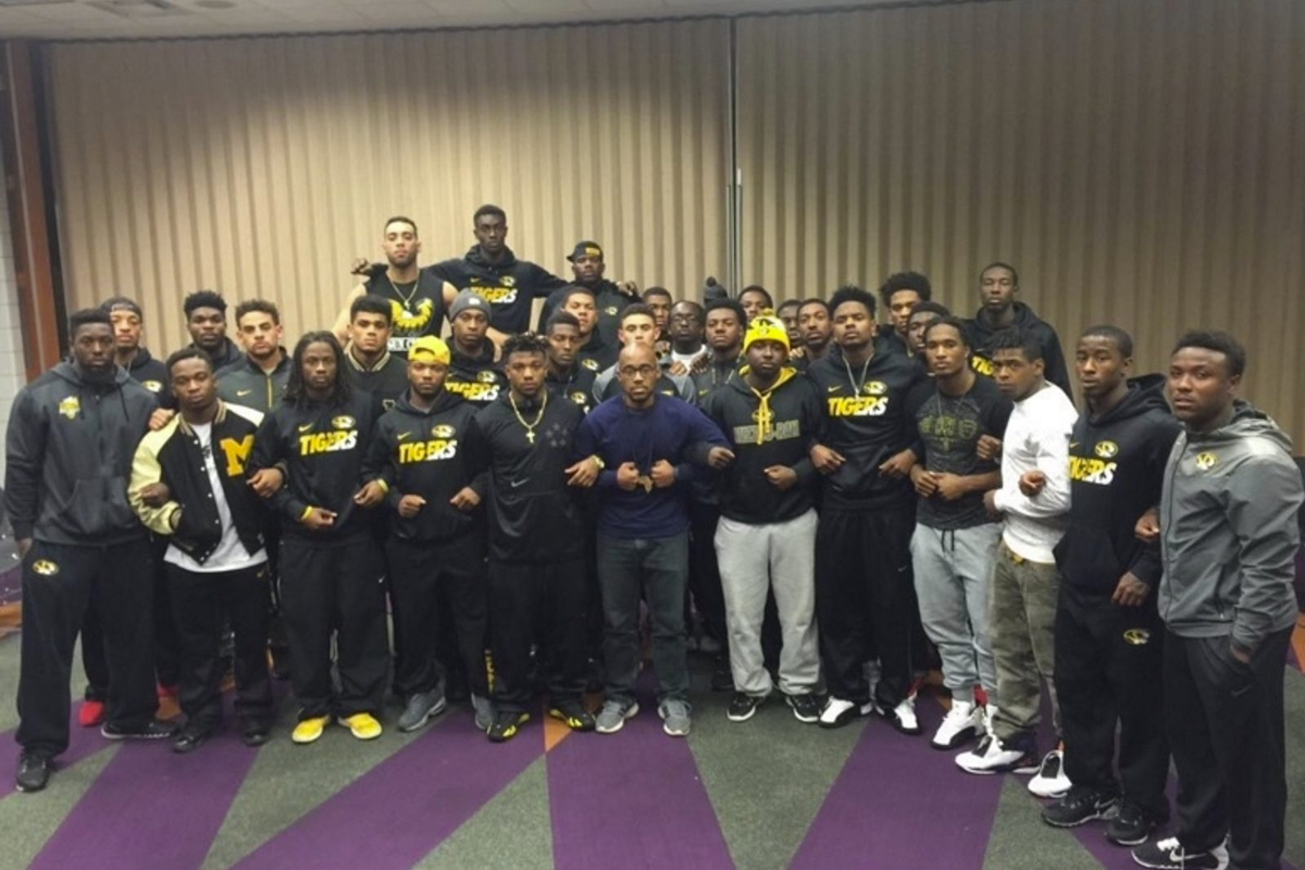 About thirty Black University of Missouri football players, many in black and gold Tigers sweatshirts, stand in rows with arms linked, alongside hunger striker Jonathan Butler at center.