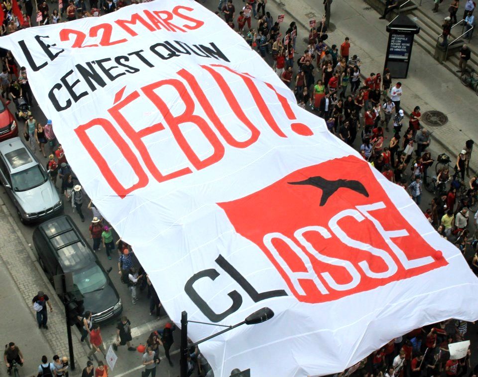 Seen from above, a giant white banner reading "Le 22 mars c'est qu'un début! CLASSE" is carried over a crowd of marchers filling a downtown street.