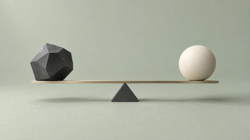 A faceted charcoal form and a smooth ivory sphere balance on a slender metal beam against a muted sage background.