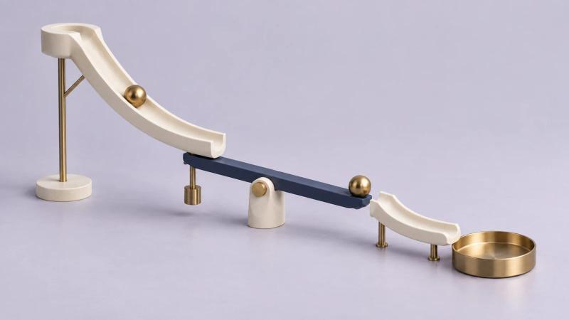 A minimal Rube Goldberg sculpture with ivory ramps, a blue lever and brass marbles on a pale lavender surface.