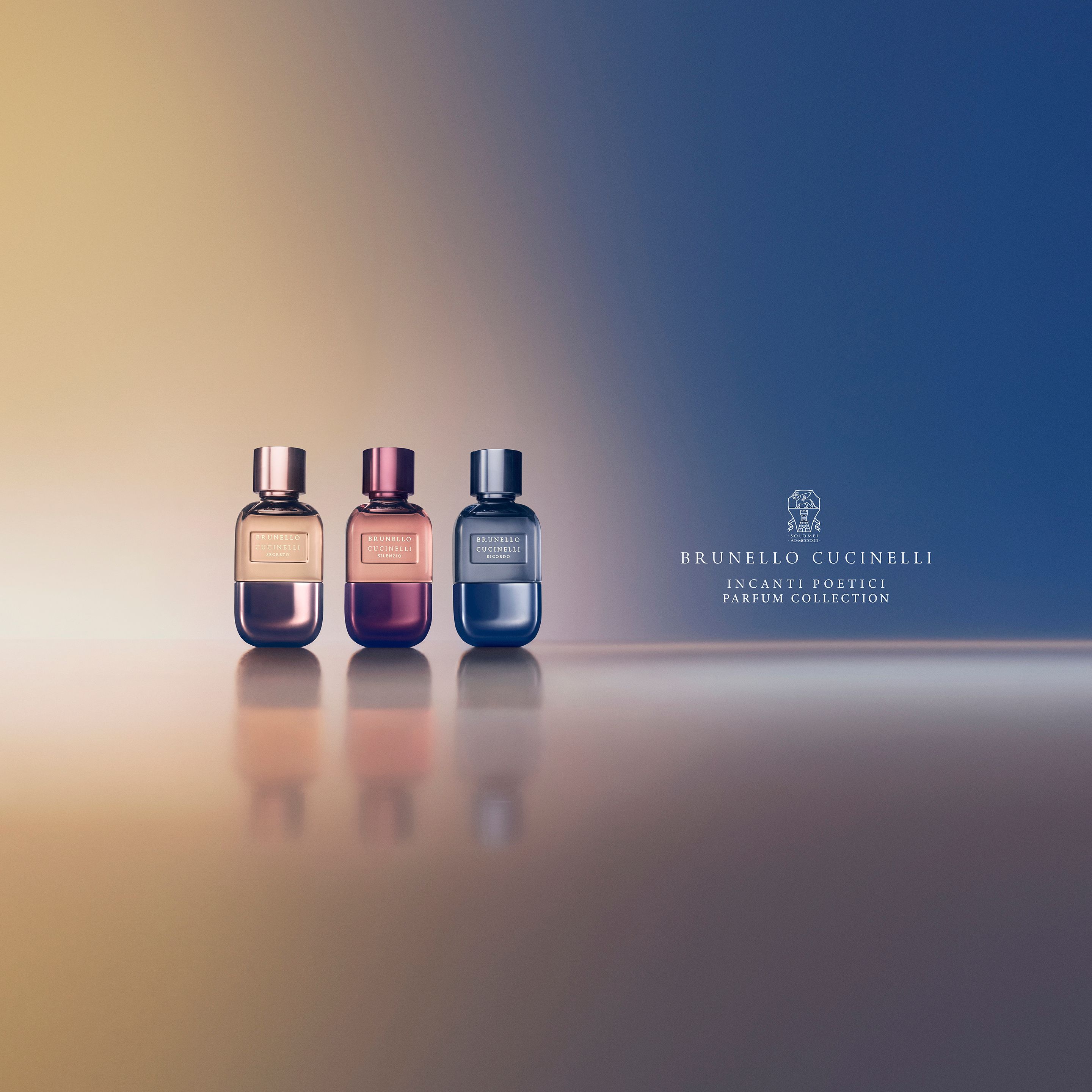 Trois flacons de parfum Brunello Cucinelli doré, rose et bleu sur une surface réfléchissante.