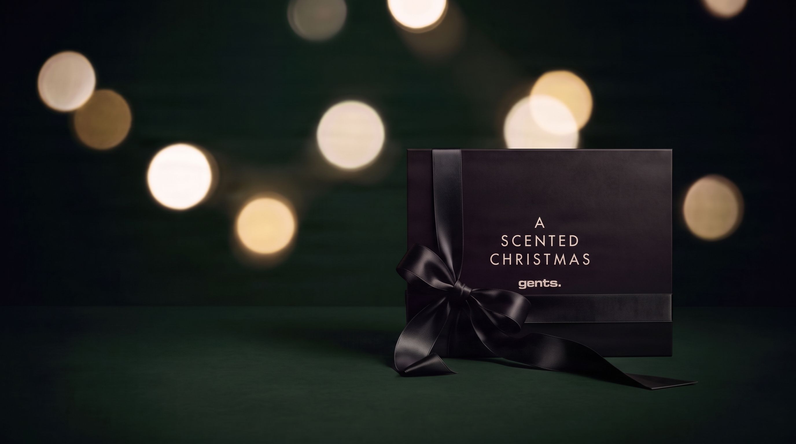 Musta lahjarasia ja rusetti tummanvihreällä taustalla, ympärillä juhlavaloja ja teksti ”A Scented Christmas”.