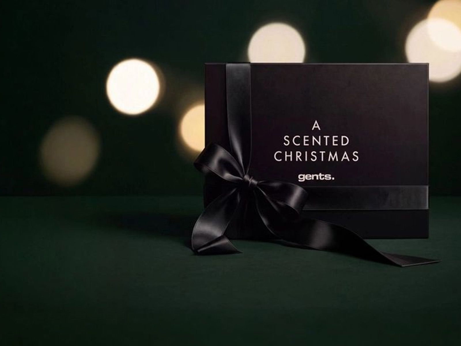 Svart presentask med svart rosett och texten ”A Scented Christmas gents” mot en mörkgrön bakgrund med suddiga julbelysningar.