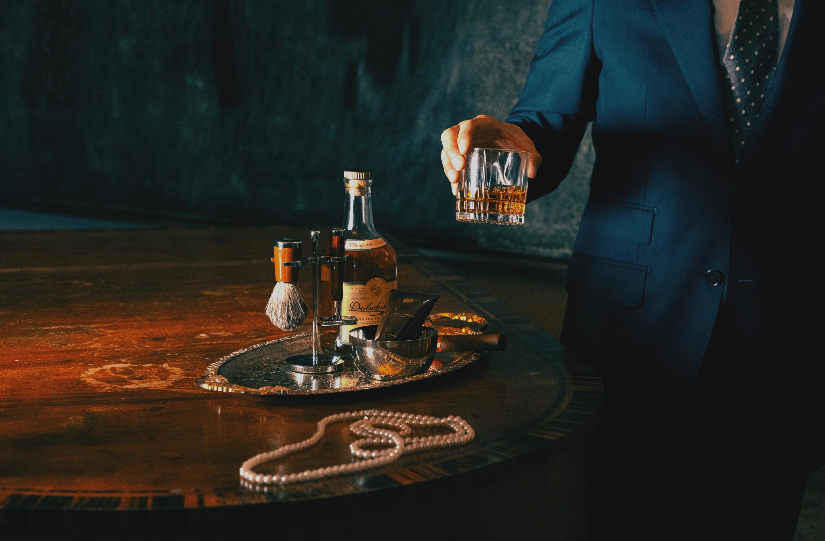En person i blå kostym håller ett glas whisky bredvid ett bord med rakset, flaska och pärlhalsband.