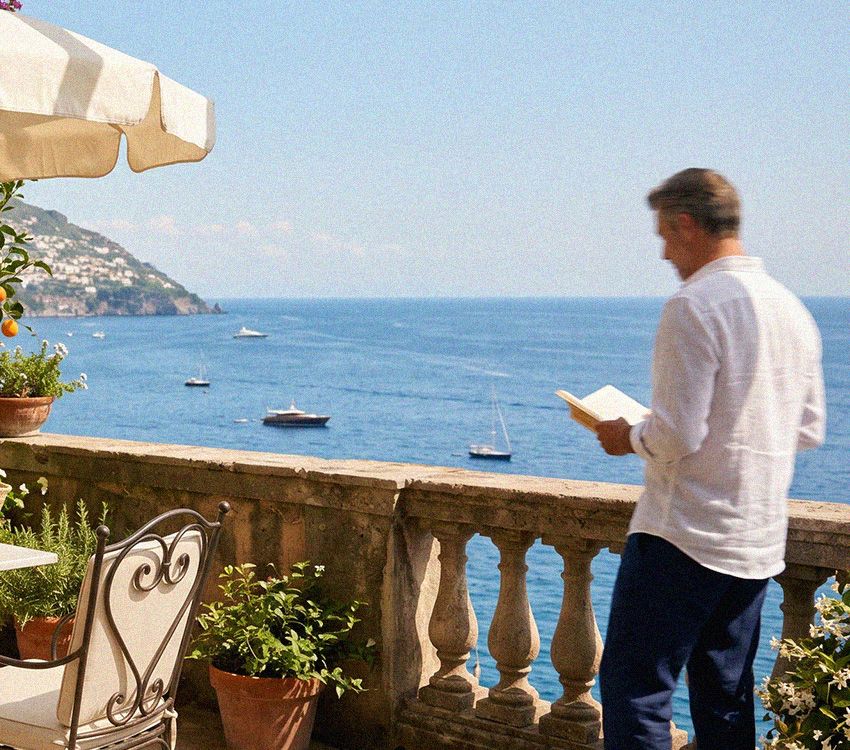 Mann sitzt auf einem Balkon und liest ein Buch, vor ihm eine ruhige blaue Meeresbucht mit einigen Booten und einer fernen Küstenstadt.