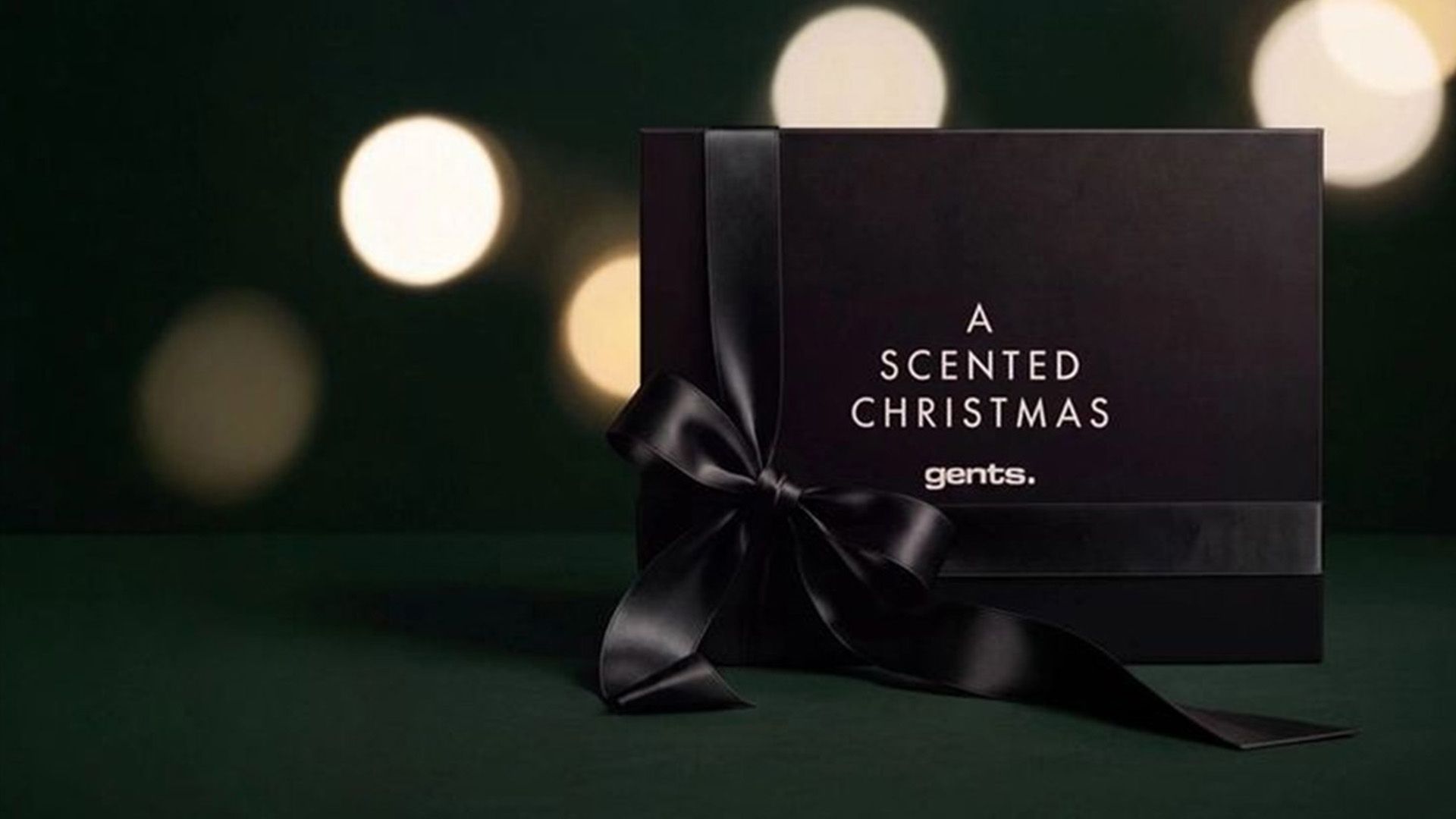 Svart presentask med svart rosett och texten ”A Scented Christmas gents” mot en mörkgrön bakgrund med suddiga julbelysningar.