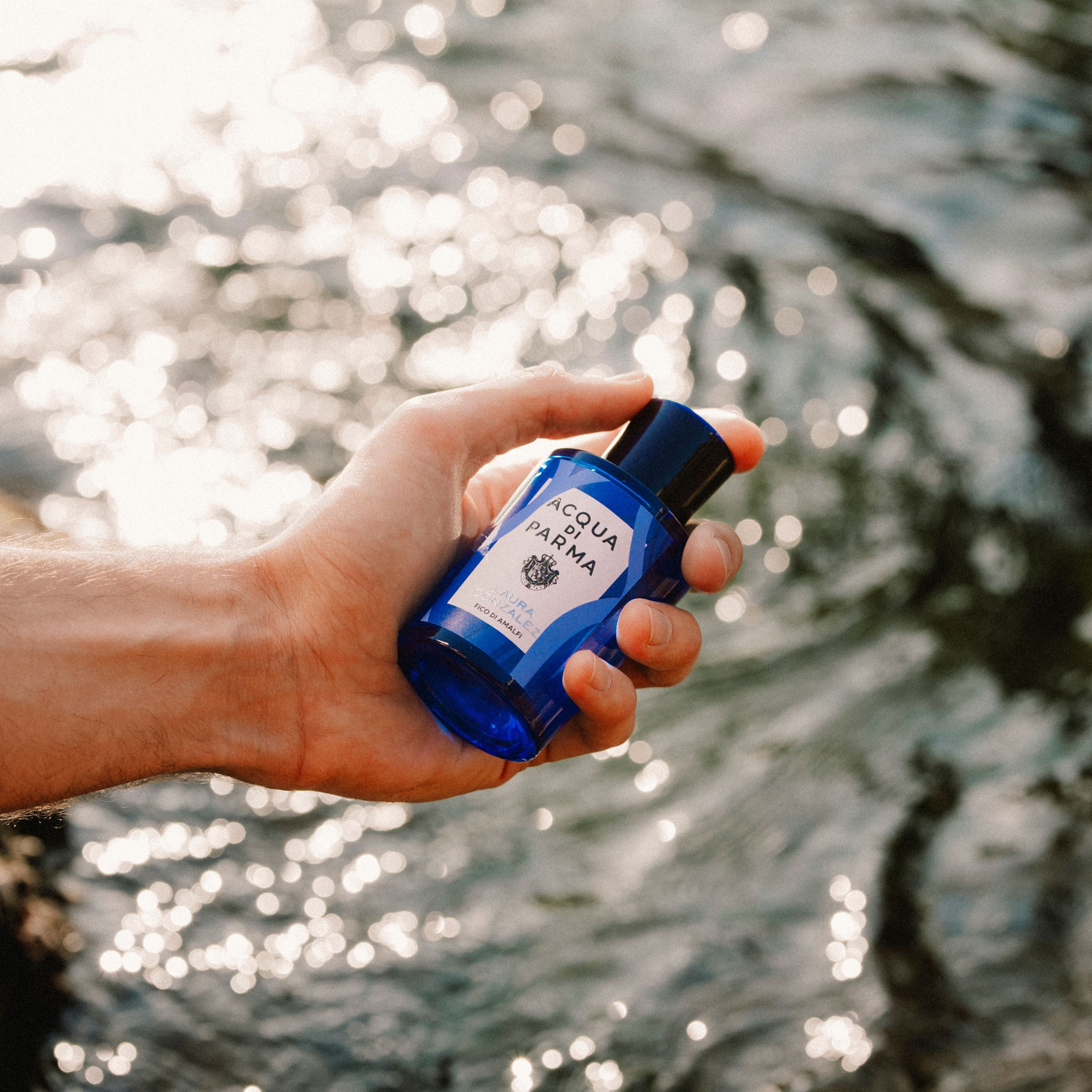Hand hält eine blaue Acqua di Parma Colonia Pura Duftflasche über funkelndem Wasser.