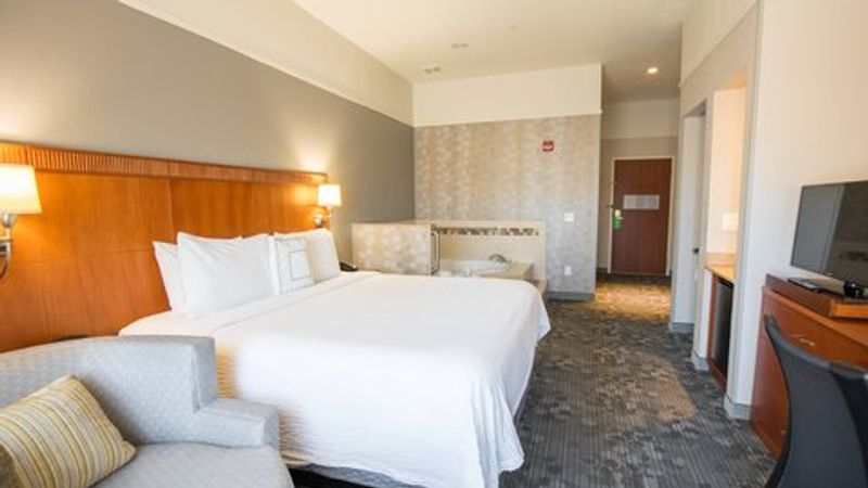 Photo of Courtyard Merced - A spacious room featuring a large bed, a private whirlpool tub, a sitting area with a chair and table, and a bathroom with a bathtub and shower.