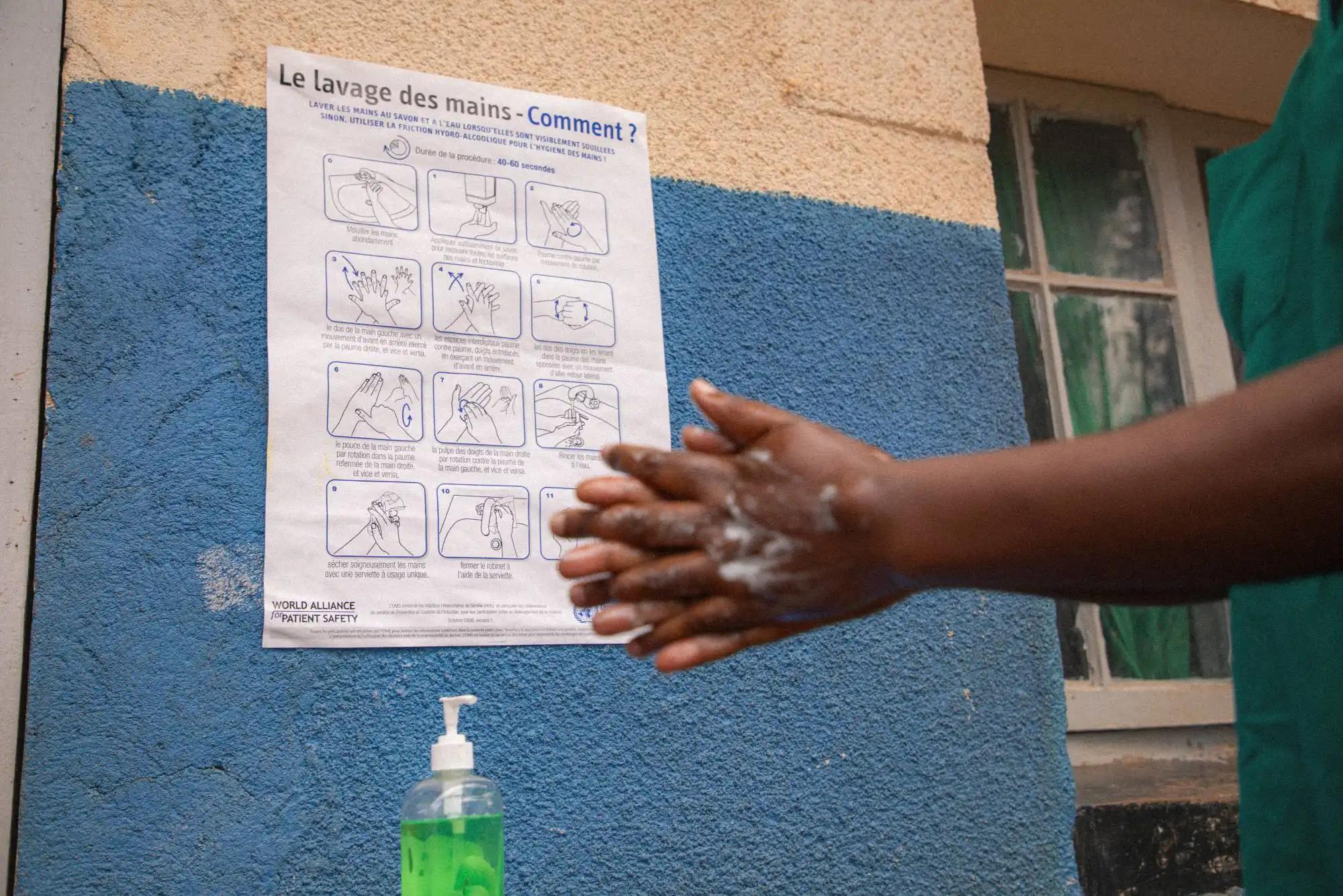 A health worker's outstretched hands, lathered in white soap, rub together beside a poster on a blue-and-cream wall titled "Le lavage des mains — Comment?" The poster illustrates the World Health Organization's handwashing technique step by step in French. Below it, a pump bottle of green liquid soap stands ready.