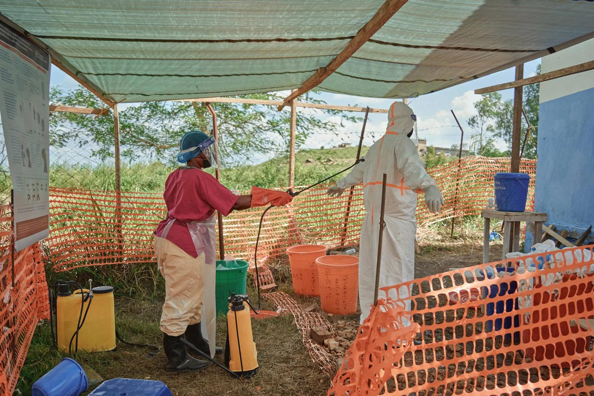 Under a tarpaulin canopy, a health worker in a face shield, pink scrubs, and orange rubber gloves sprays disinfectant from a hand pump onto a colleague in a white protective suit, who stands with arms outstretched. Around them are orange mesh fencing, buckets labeled with chlorine concentrations, and yellow sprayer tanks.