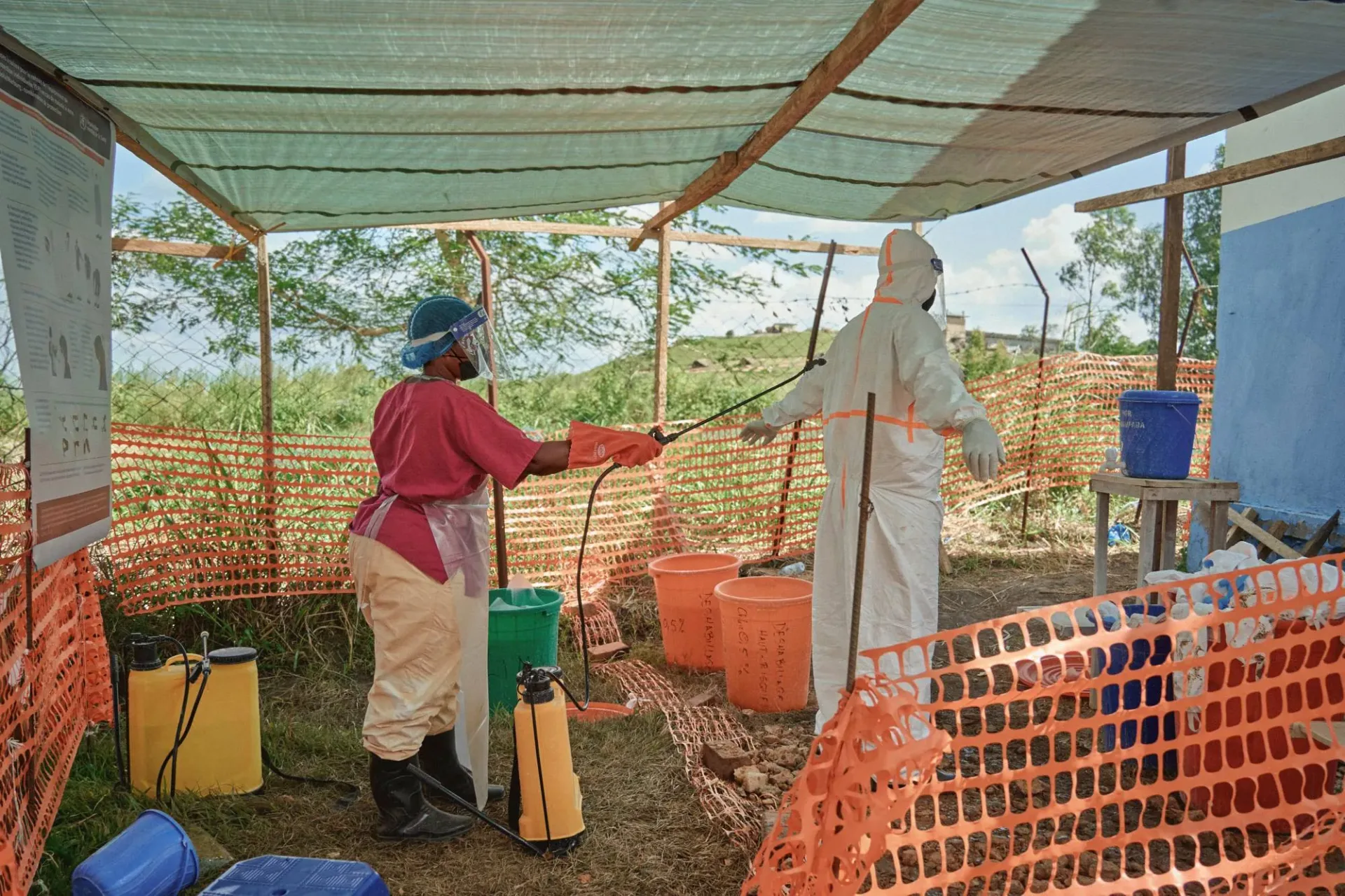Under a tarpaulin canopy, a health worker in a face shield, pink scrubs, and orange rubber gloves sprays disinfectant from a hand pump onto a colleague in a white protective suit, who stands with arms outstretched. Around them are orange mesh fencing, buckets labeled with chlorine concentrations, and yellow sprayer tanks.