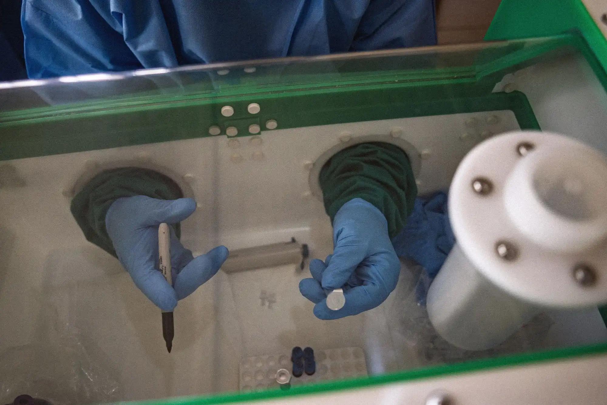 Seen from above, a lab worker in a blue gown reaches through the sealed glove ports of a green-framed glovebox, blue nitrile gloves over dark green sleeves. One hand holds a black marker ready to write; the other holds a small round vial cap. Inside the chamber, trays of sample tubes and dark vial caps sit on the white work surface.