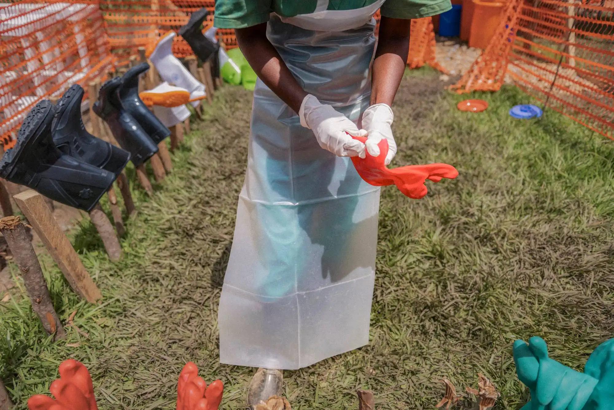 A health worker in a clear plastic apron over green scrubs pulls a heavy red rubber glove on over white medical gloves. Around them, rubber boots dry upside down on wooden stakes and pairs of red and green gloves are laid out on the grass, inside an enclosure of orange mesh fencing.