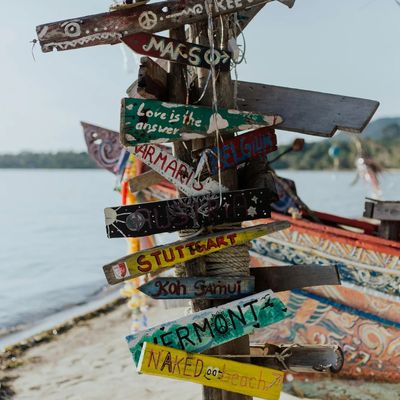 A rustic wooden signpost on a tropical beach with multiple hand-painted directional signs pointing to various destinations such as 'Koh Samui,' 'Stuttgart,' 'Belgium,' and 'Naked Beach.' The signs are colorful and weathered, attached to a wooden pole with ropes and surrounded by a traditional wooden boat and ocean view in the background.