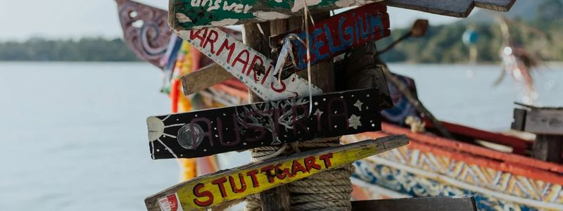 A rustic wooden signpost on a tropical beach with multiple hand-painted directional signs pointing to various destinations such as 'Koh Samui,' 'Stuttgart,' 'Belgium,' and 'Naked Beach.' The signs are colorful and weathered, attached to a wooden pole with ropes and surrounded by a traditional wooden boat and ocean view in the background.