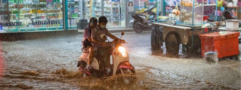 A motorbike struggles through a flooded street in Bangkok during heavy rain, with street food stalls and a pharmacy in the background – a reminder of Thailand’s intense rainy season.