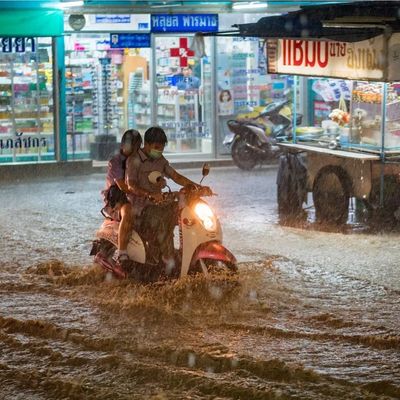 A motorbike struggles through a flooded street in Bangkok during heavy rain, with street food stalls and a pharmacy in the background – a reminder of Thailand’s intense rainy season.