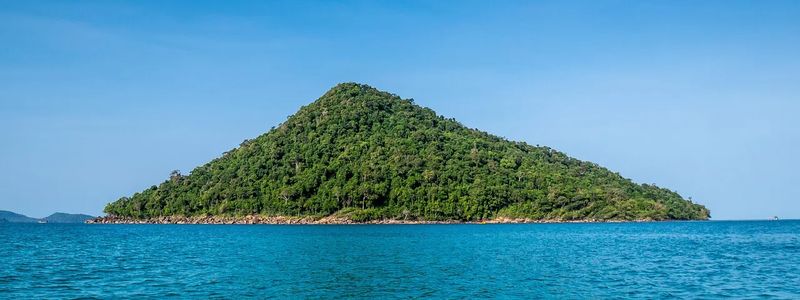 A lush green island surrounded by turquoise waters under a clear blue sky in Cambodia, showcasing the serene beauty of Koh Rong or Koh Rong Samloem.